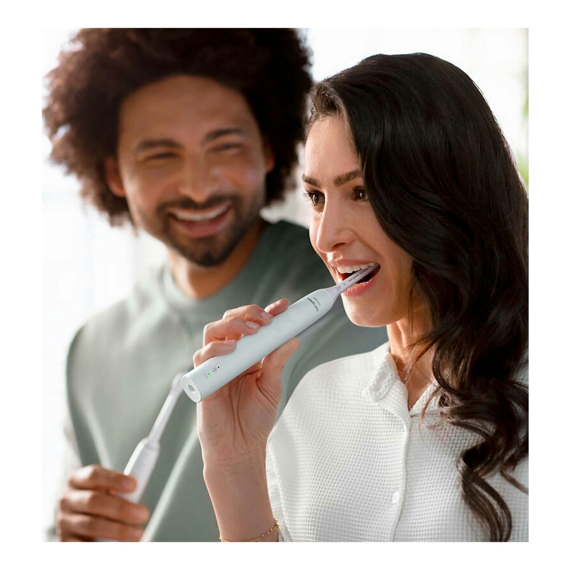 Vista frontal de una mujer cepillándose con un cepillo dental eléctrico Philips blanco; hombre desenfocado detrás.