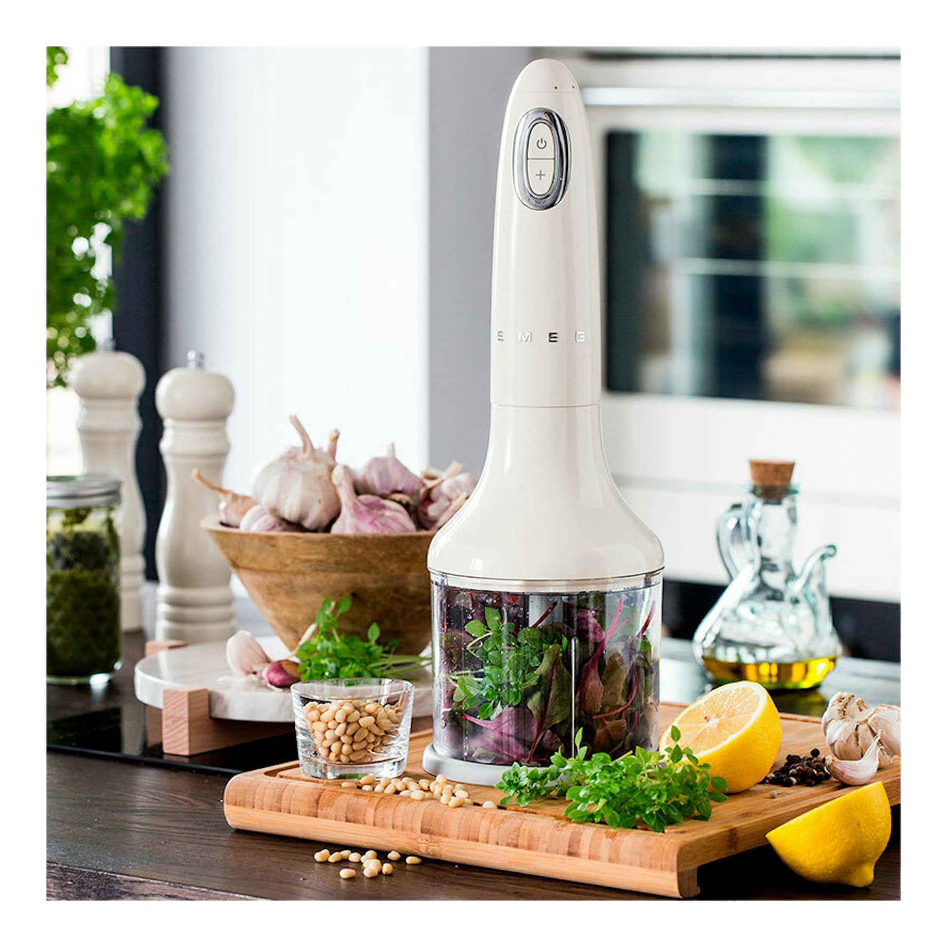 Batidora de mano Smeg HBF03CREU crema, vista frontal, con accesorio picador y alimentos en una encimera.