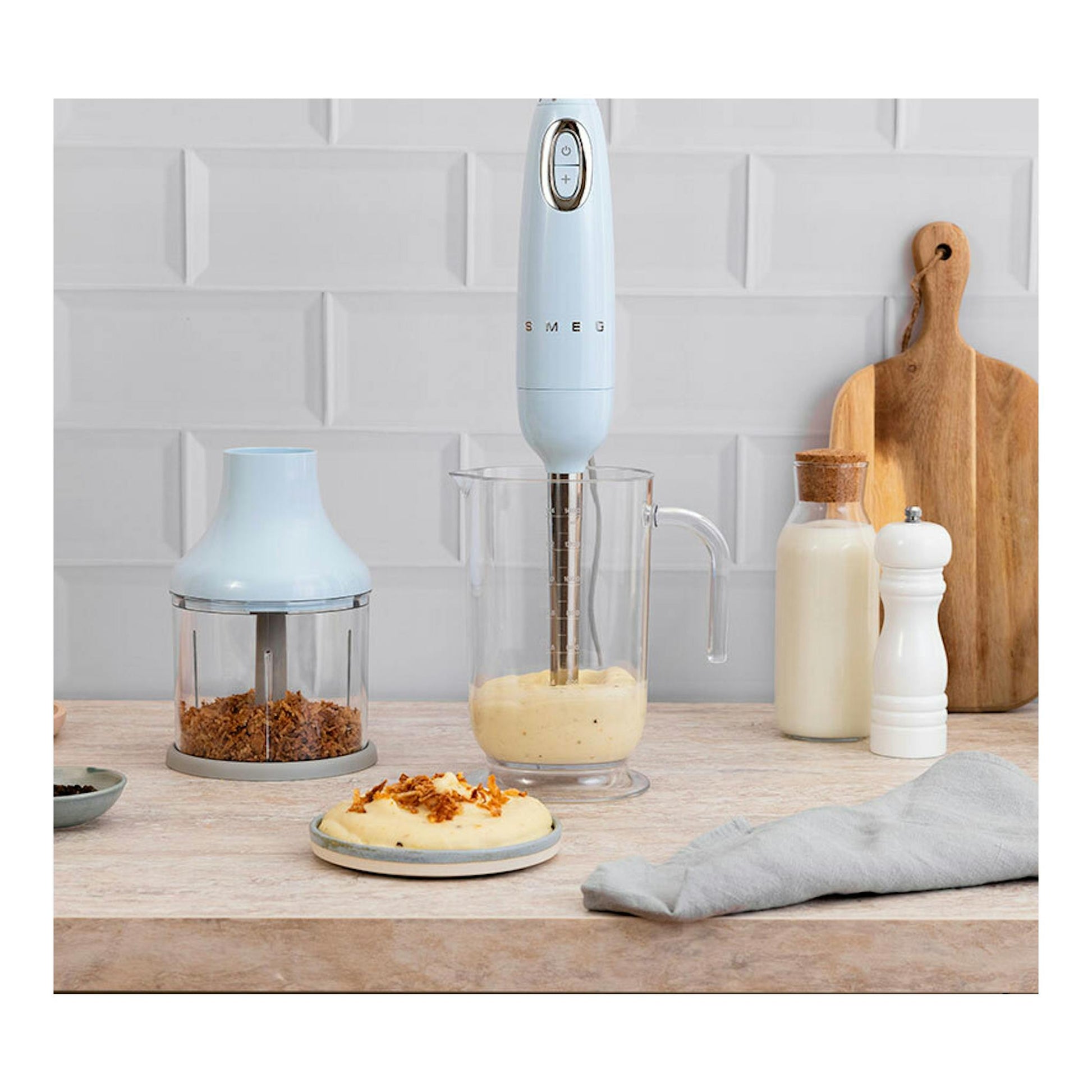 Batidora de mano Smeg HBF03PBEU azul pastel, vista frontal, en vaso medidor sobre encimera con accesorios.