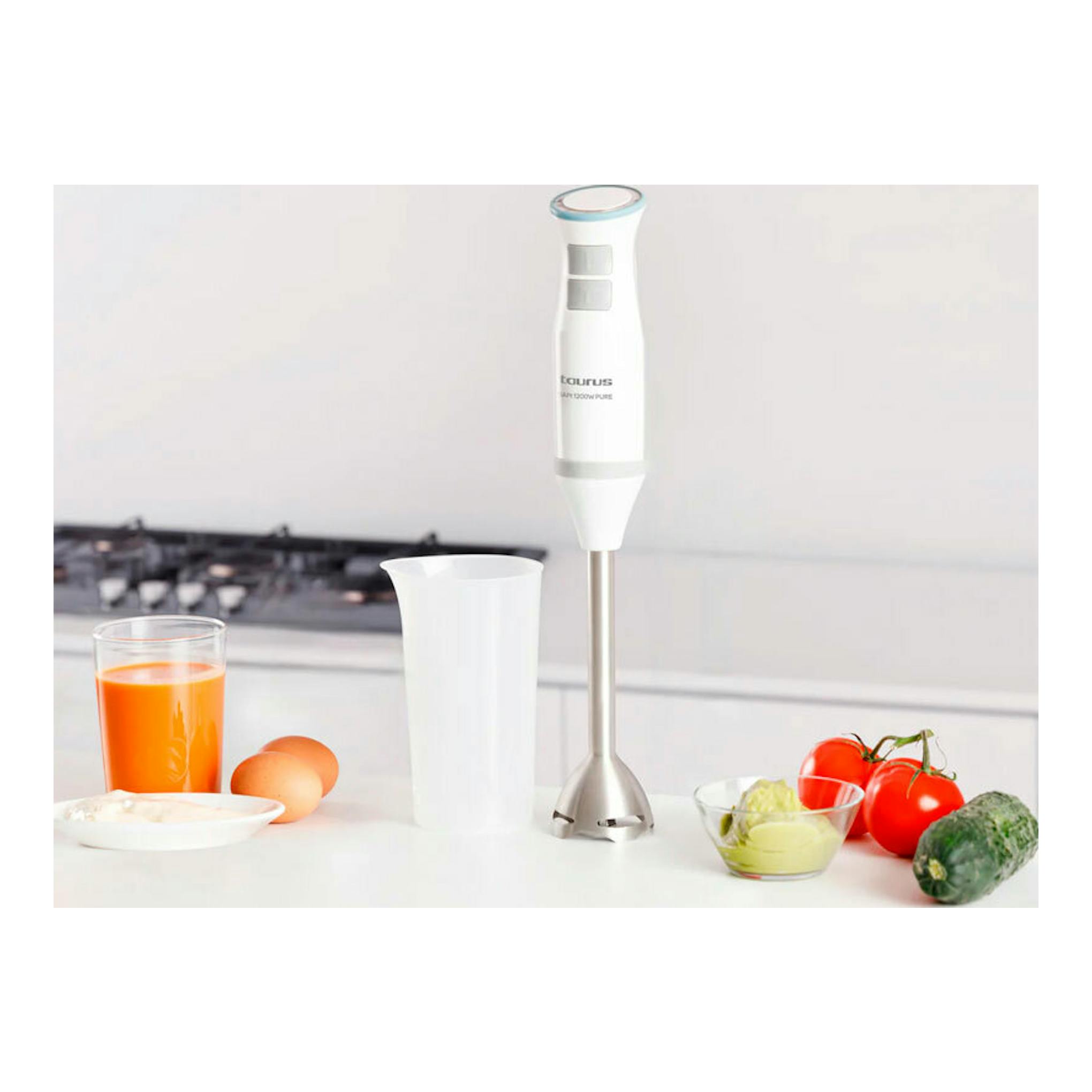 Batidora de mano Taurus Bapi 1200 Pure blanca, vista frontal, con vaso y alimentos sobre una encimera de cocina.