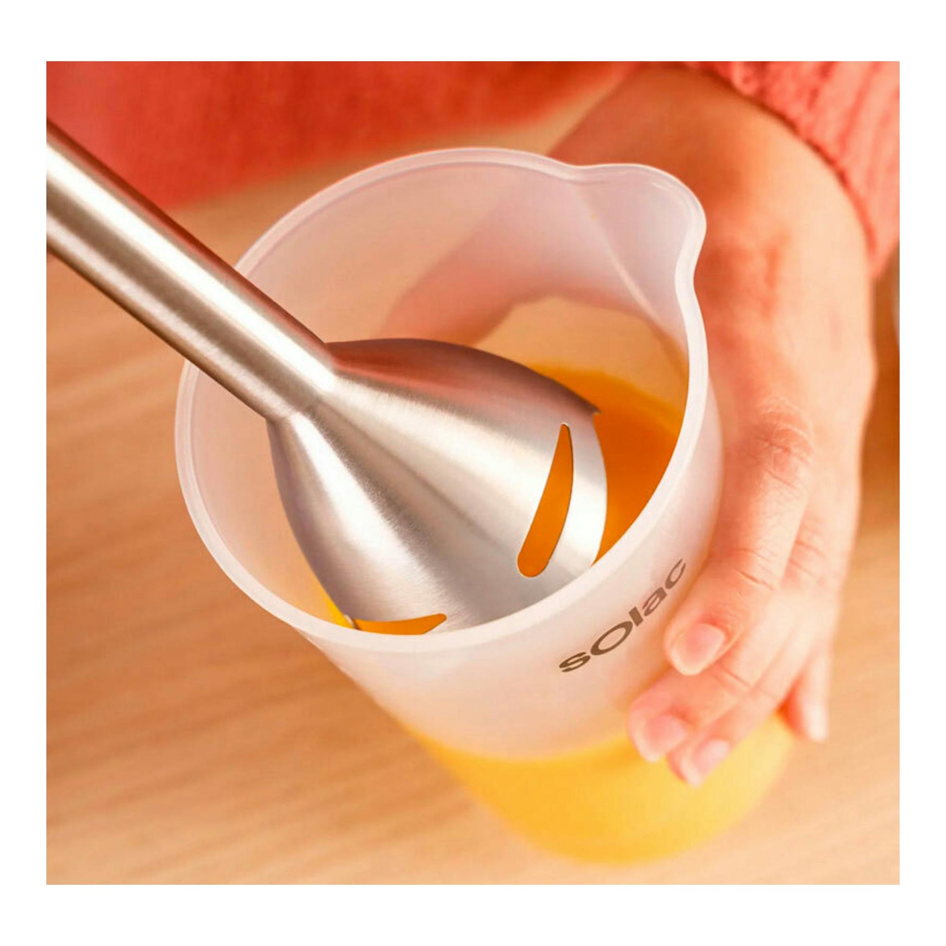 Batidora de inmersión Solac BA5617 en primer plano, batiendo líquido naranja en un vaso medidor.
