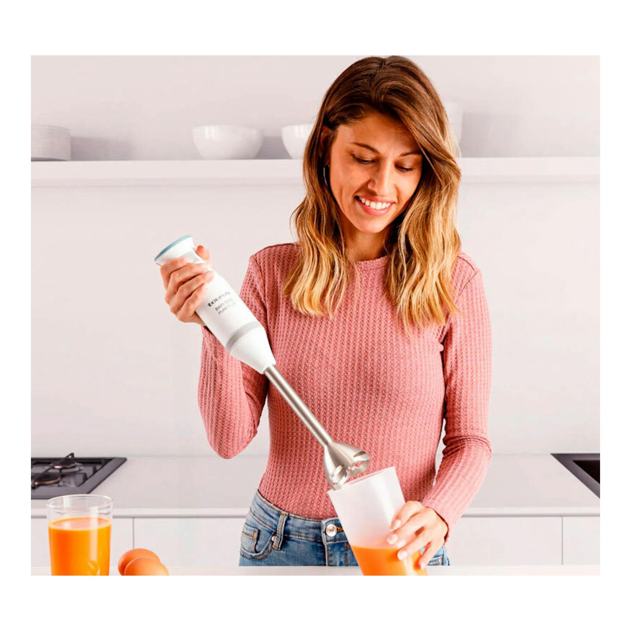 Vista frontal de una mujer usando una batidora de mano Taurus Bapi 1200 Pure Plus en cocina, con líquido naranja.