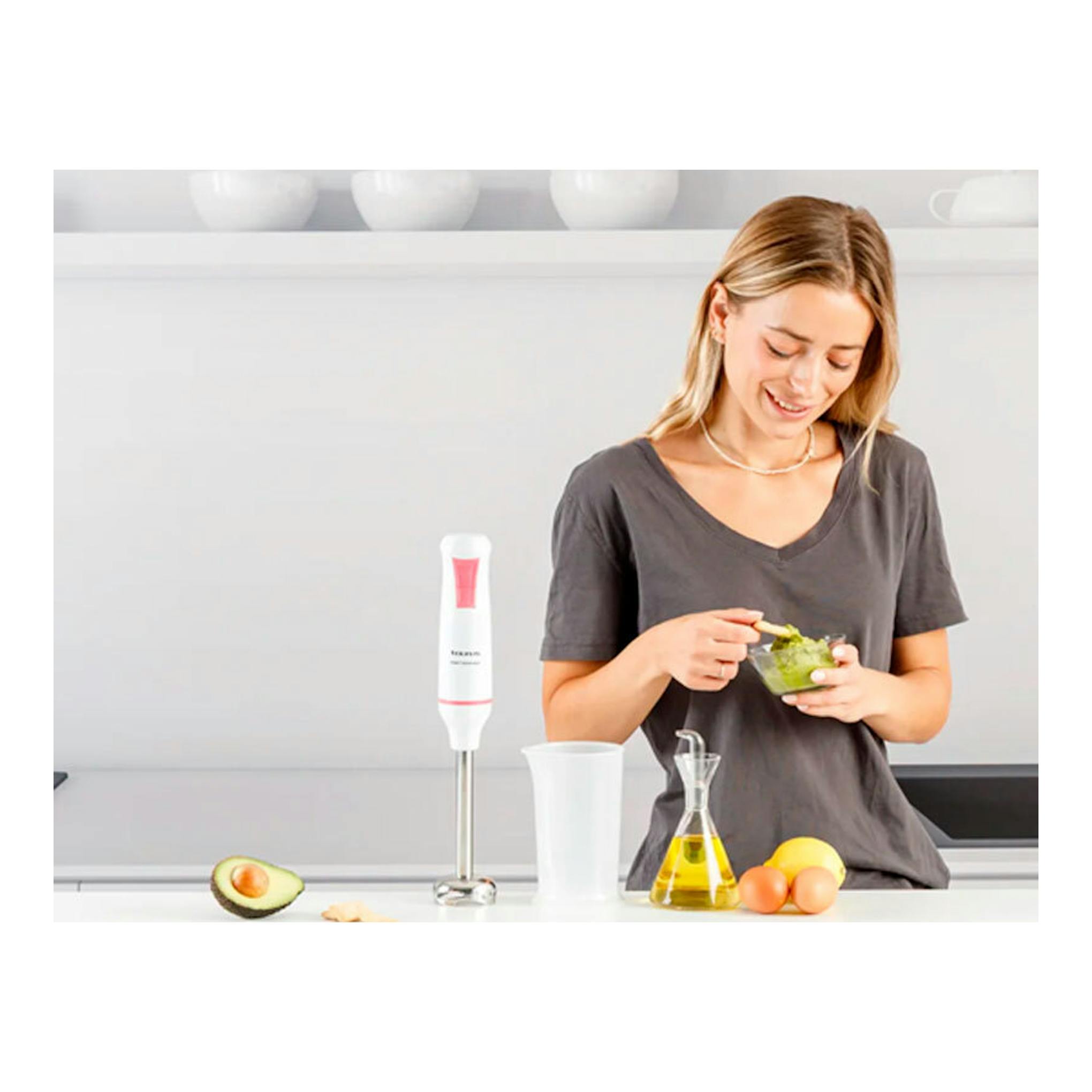 Batidora de mano Taurus Robot 1000 Inox blanca con detalle rosa sobre encimera, junto a una mujer preparando comida.