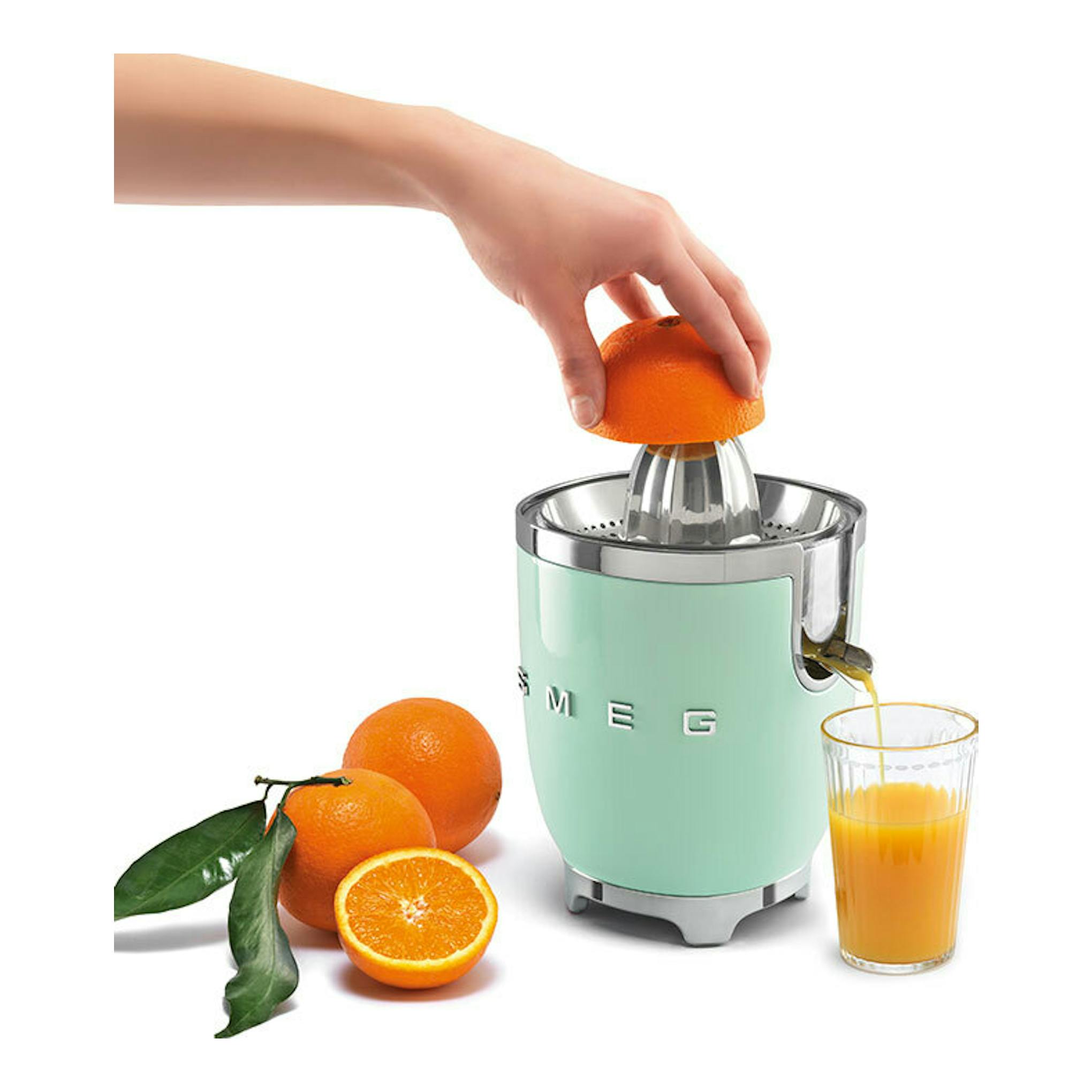 Exprimidor Smeg CJF11PGEU verde menta, vista frontal, con una mano exprimiendo una naranja y un vaso de zumo.