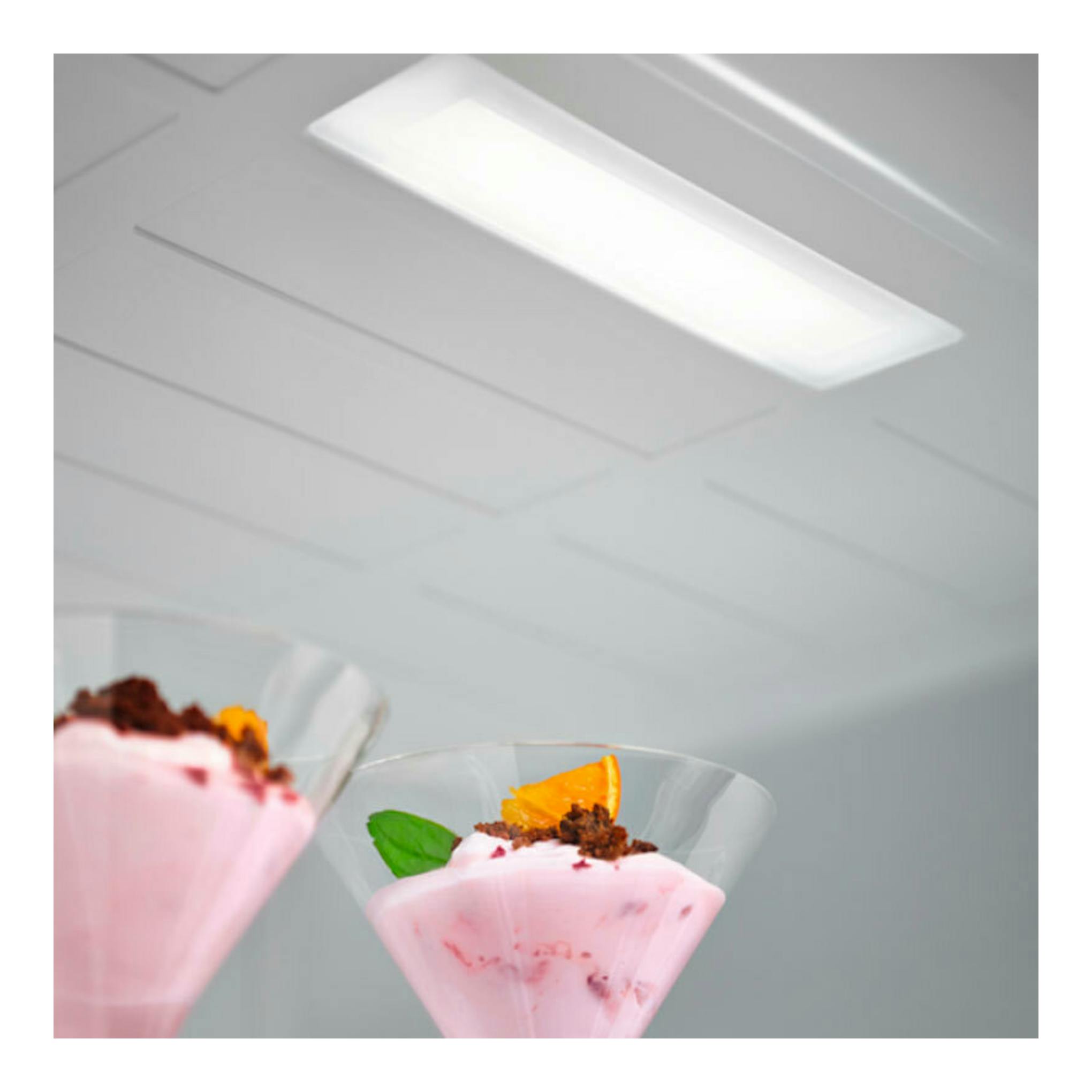 Interior de frigorífico blanco con luz LED y dos copas de postre rosa en primer plano.