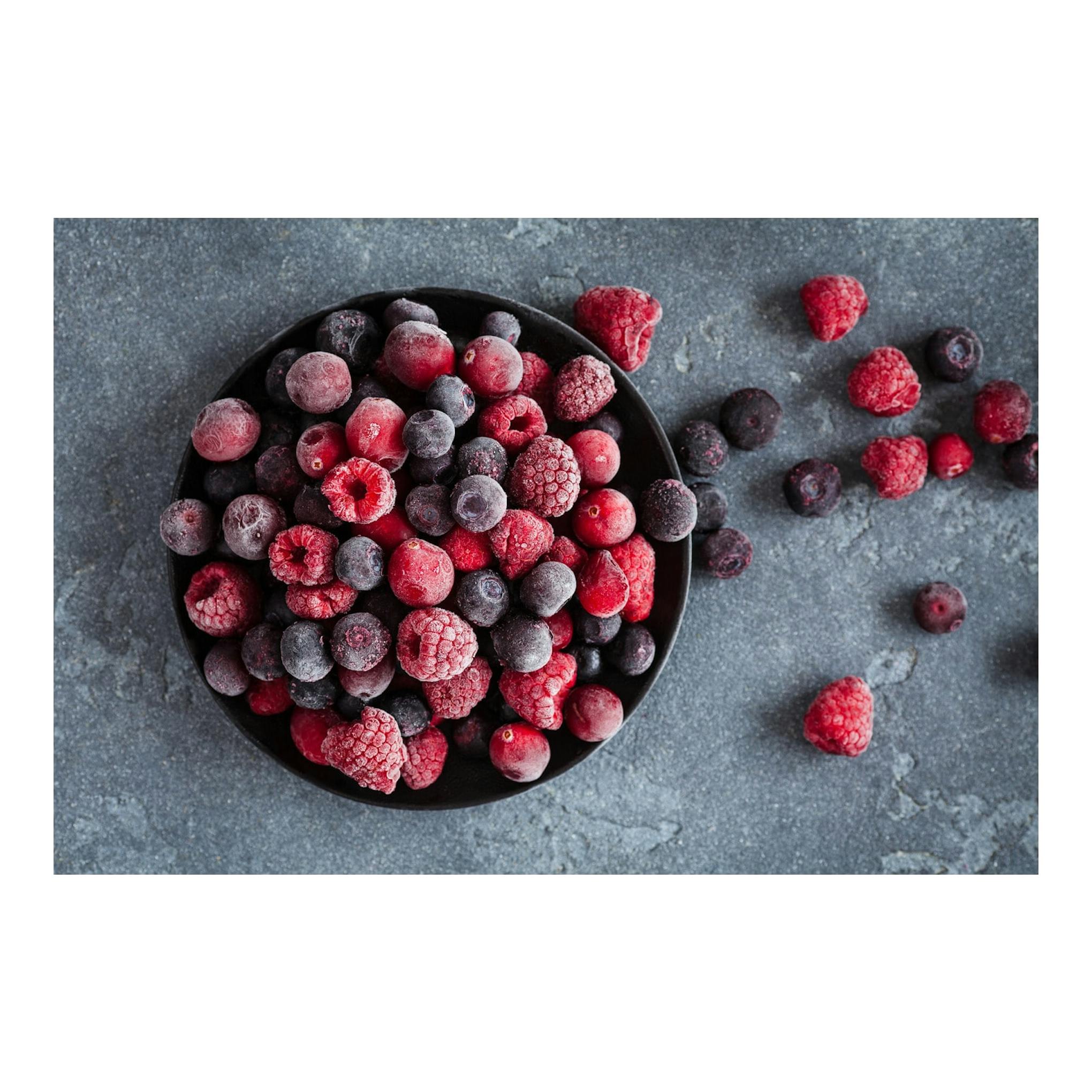 Vista cenital de un cuenco negro con frutos rojos y arándanos congelados sobre fondo gris.