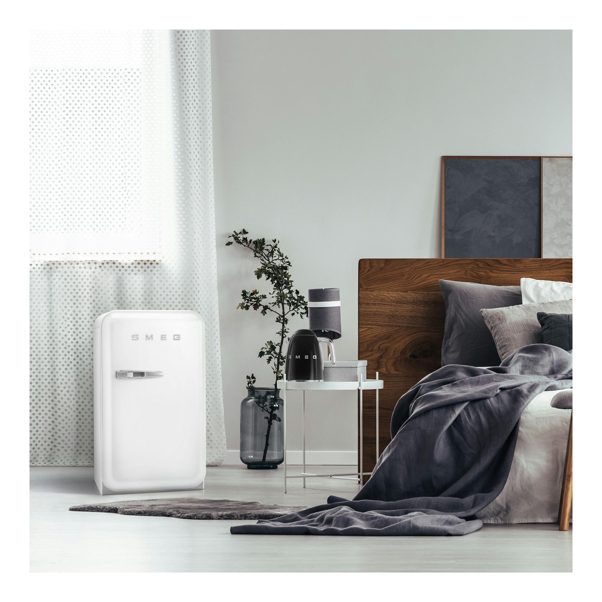 Frigorífico compacto Smeg FAB5RWH5 blanco, vista frontal, junto a una cama en un dormitorio.