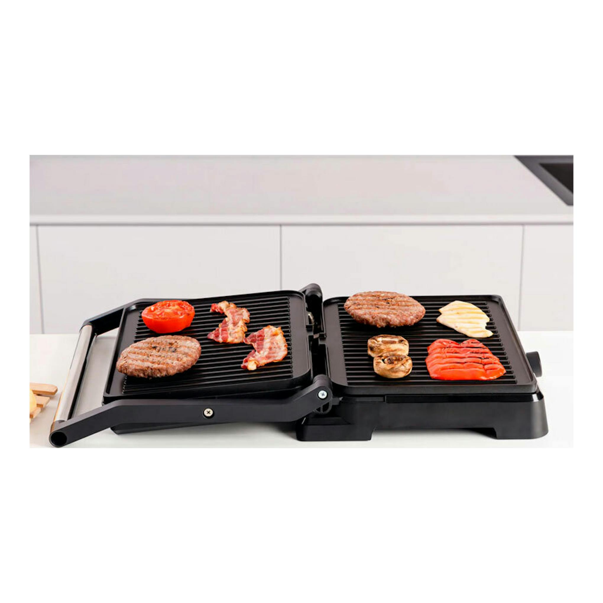 Grill Taurus Bistro negra, vista frontal, abierta sobre encimera blanca, con alimentos en ambas planchas.