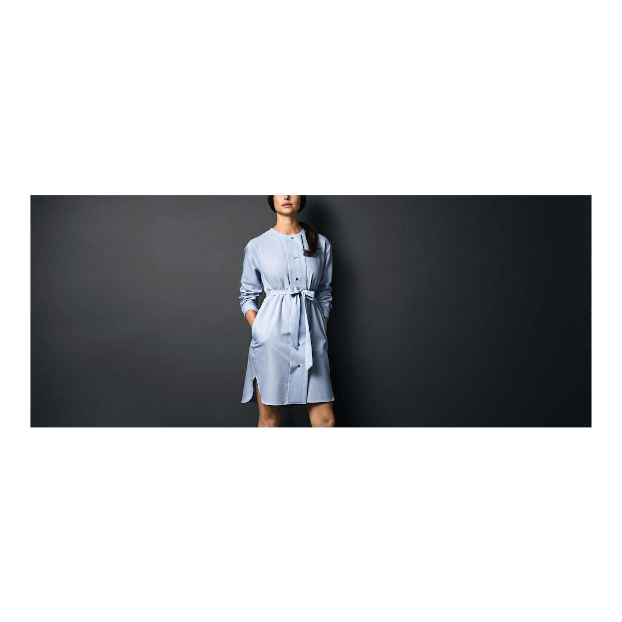Mujer de pie con vestido camisero azul claro y cinturón, sobre fondo gris oscuro, vista frontal recortada.