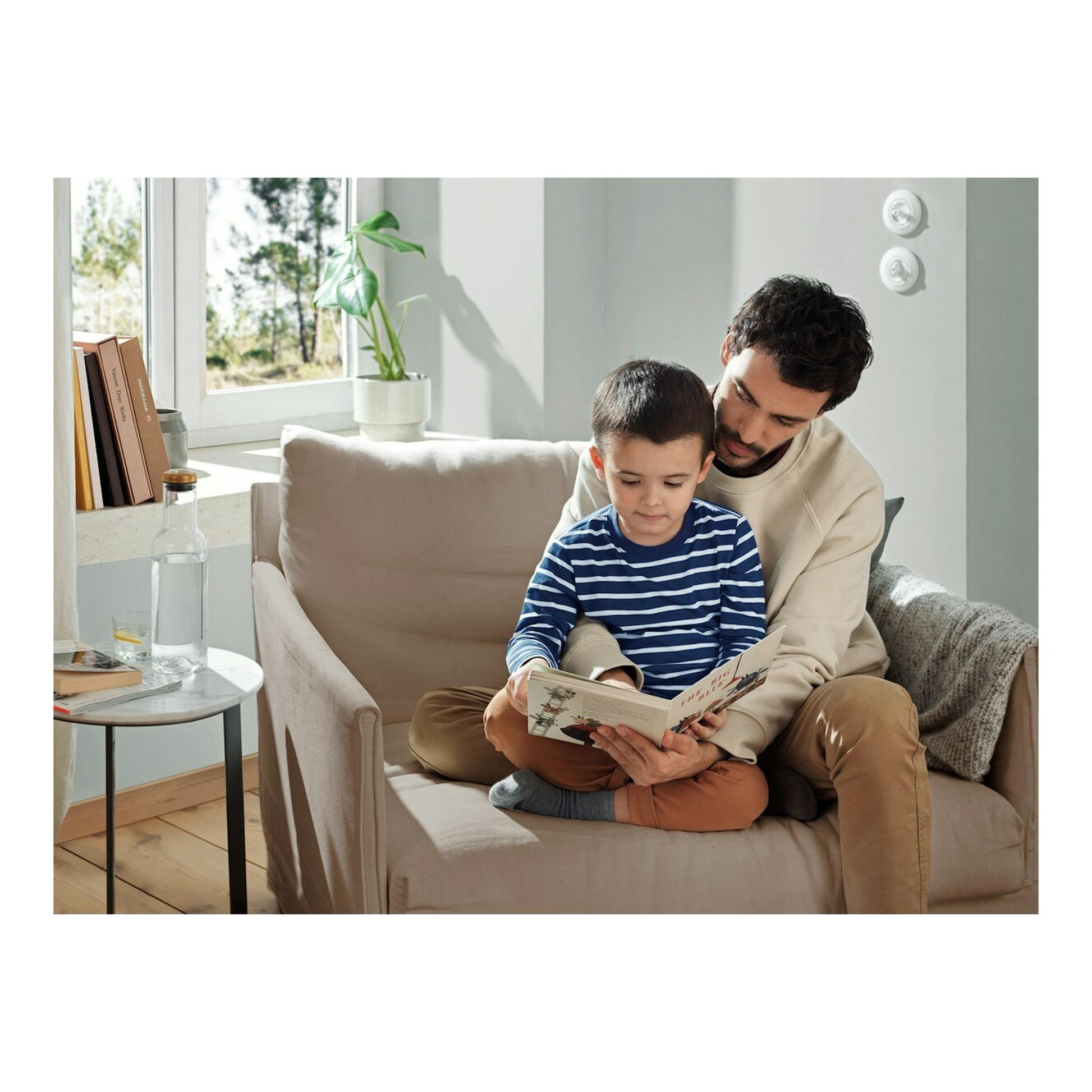 Padre e hijo sentados en un sofá leyendo un libro, en una sala iluminada junto a una ventana.