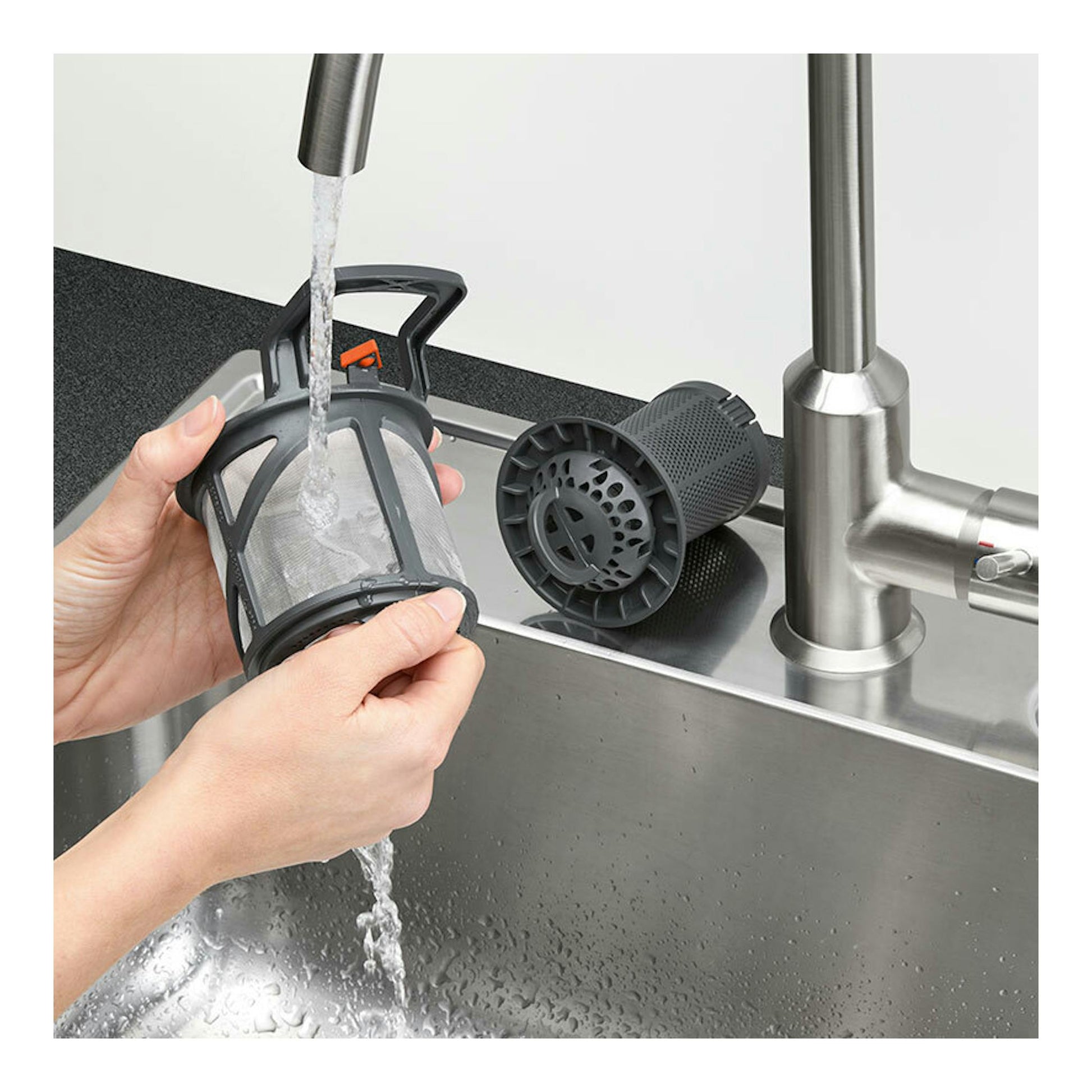 Manos enjuagando el filtro de un lavavajillas bajo el grifo, junto a un fregadero de acero inoxidable.