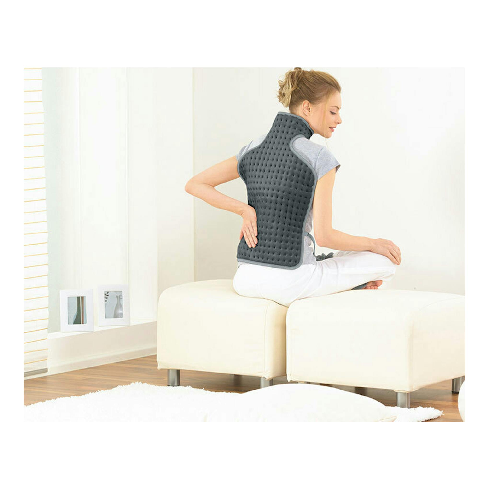 Manta eléctrica Beurer HK-53 gris colocada en la espalda de una mujer sentada de perfil sobre un asiento blanco.
