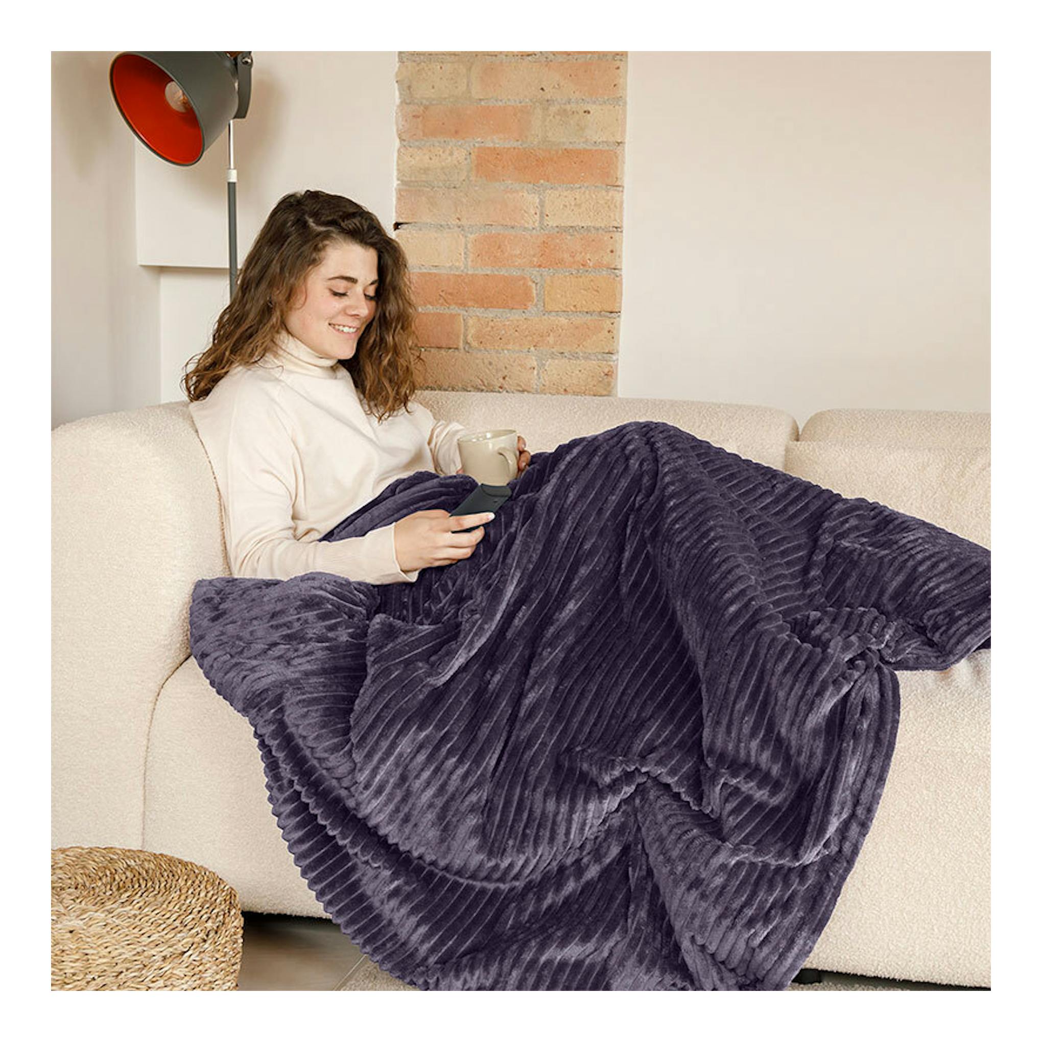 Manta eléctrica Solac CT8706 morada sobre sofá, con mujer sentada y mando en la mano.