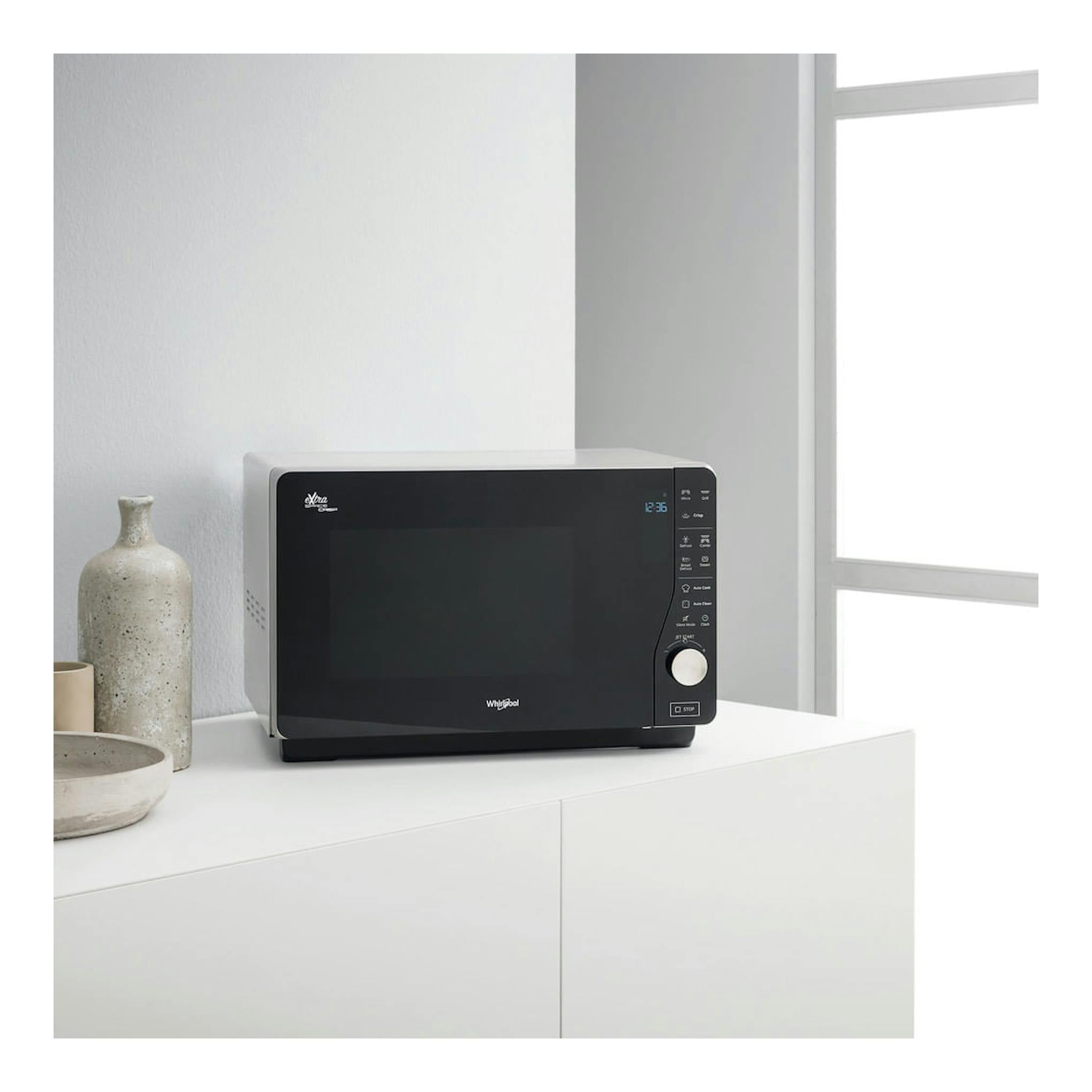 Microondas Whirlpool MWF 427 SL negro y plateado, vista frontal sobre mueble blanco junto a una ventana.