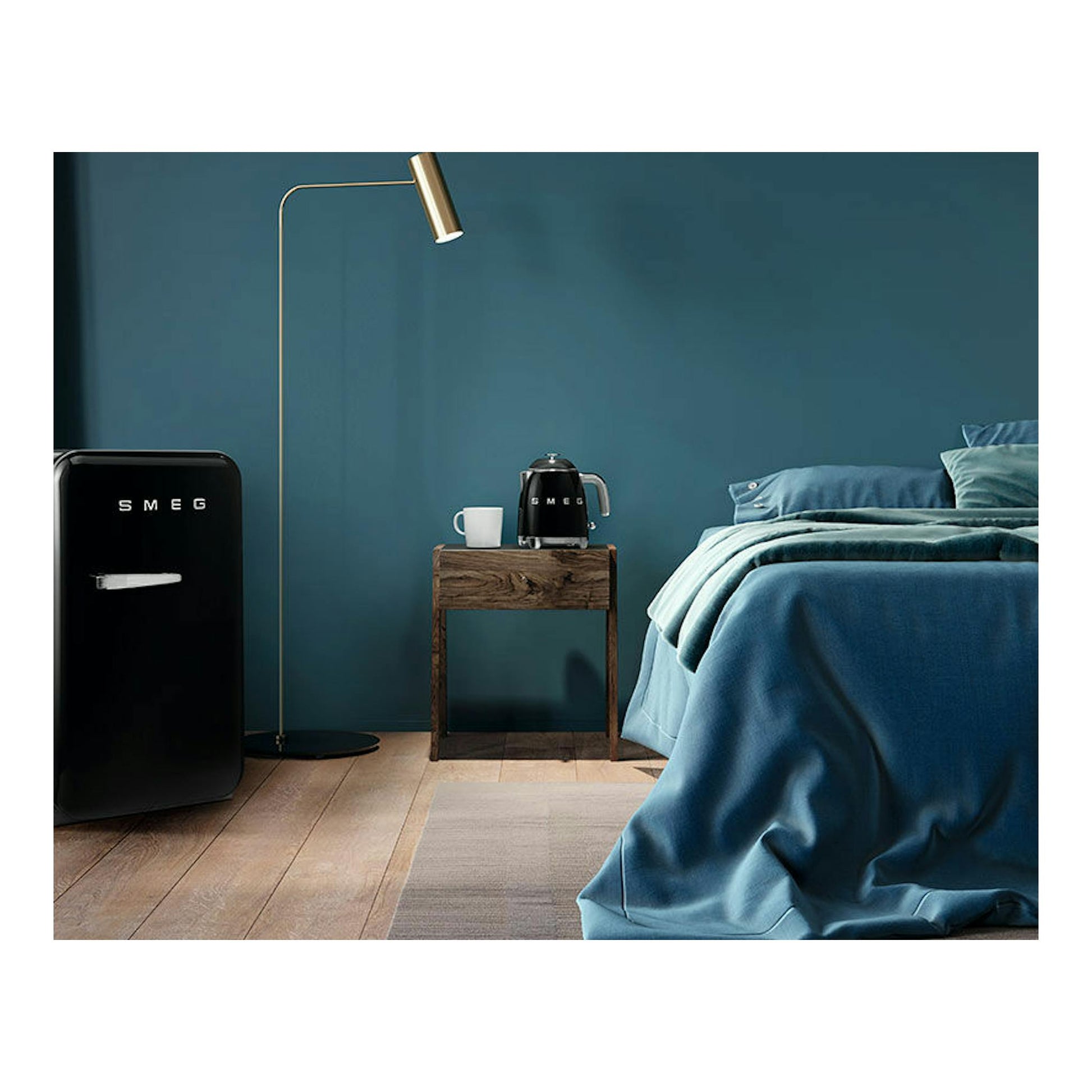Mini hervidor Smeg KLF05BLEU negro, vista frontal, sobre mesita junto a una taza en un dormitorio azul.