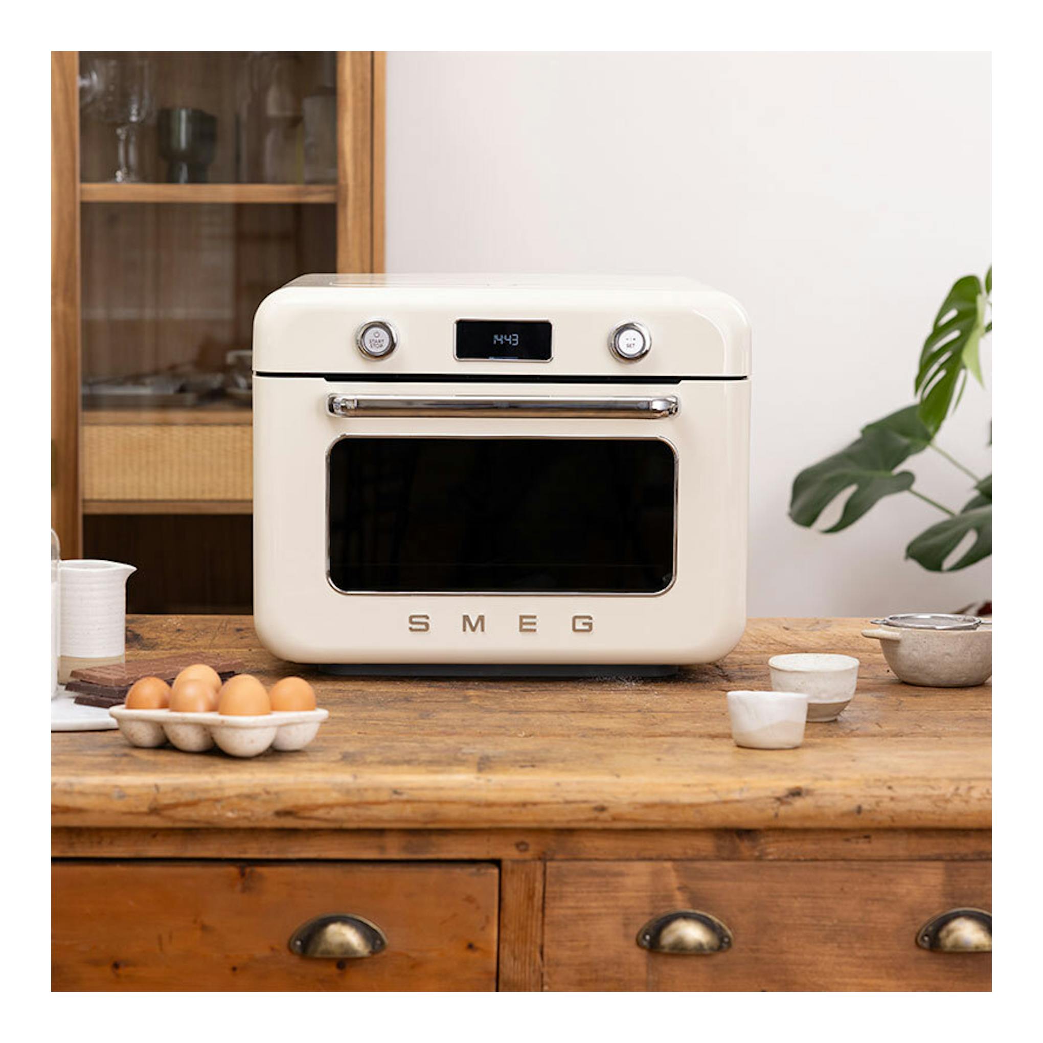 Minihorno Smeg COF01CREU crema, vista frontal con puerta negra y display digital sobre mesa de madera.