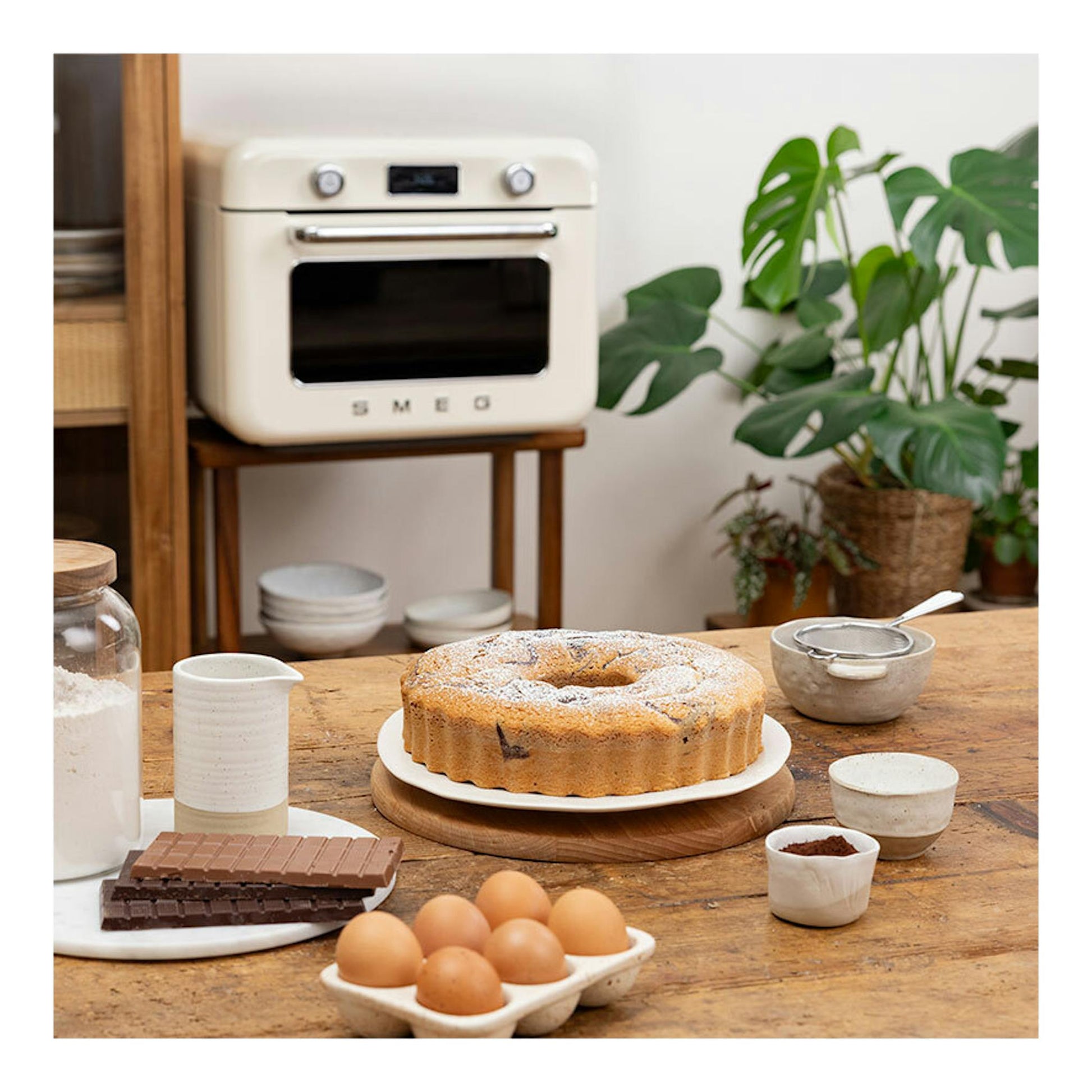 Minihorno Smeg COF01WHEU blanco, vista frontal, en una cocina con bizcocho, huevos y chocolate sobre mesa.