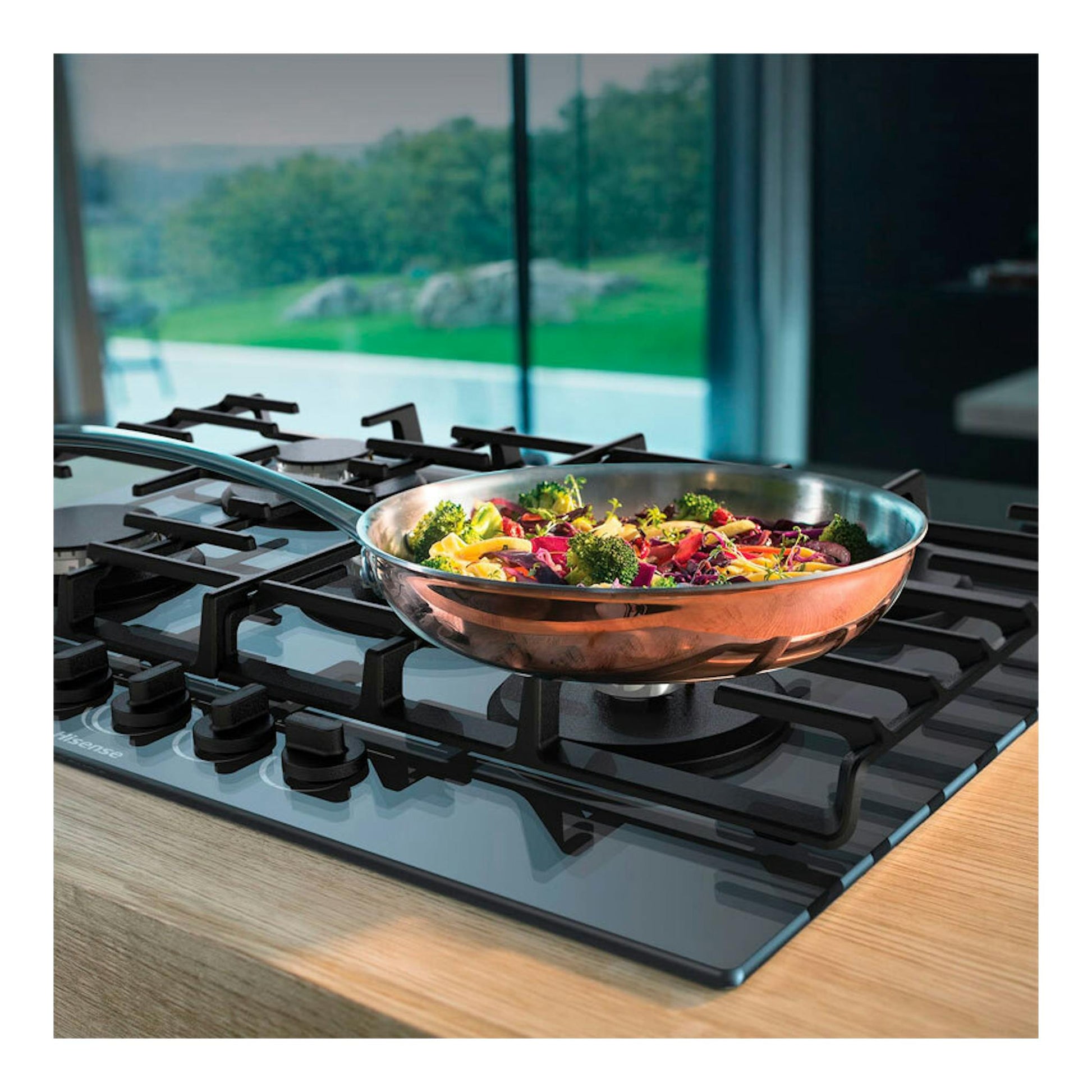 Placa de gas de cristal Hisense GG773B, vista en ángulo, con sartén de cobre y verduras sobre parrillas negras.