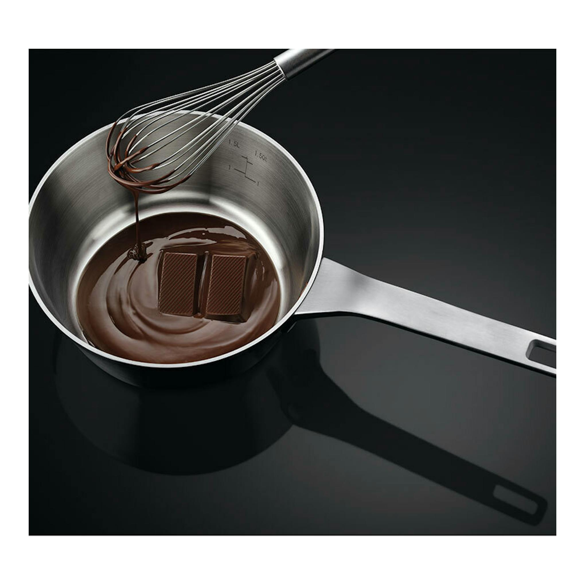 Vista cenital de una placa de inducción negra con un cazo de acero, varillas y chocolate fundido.
