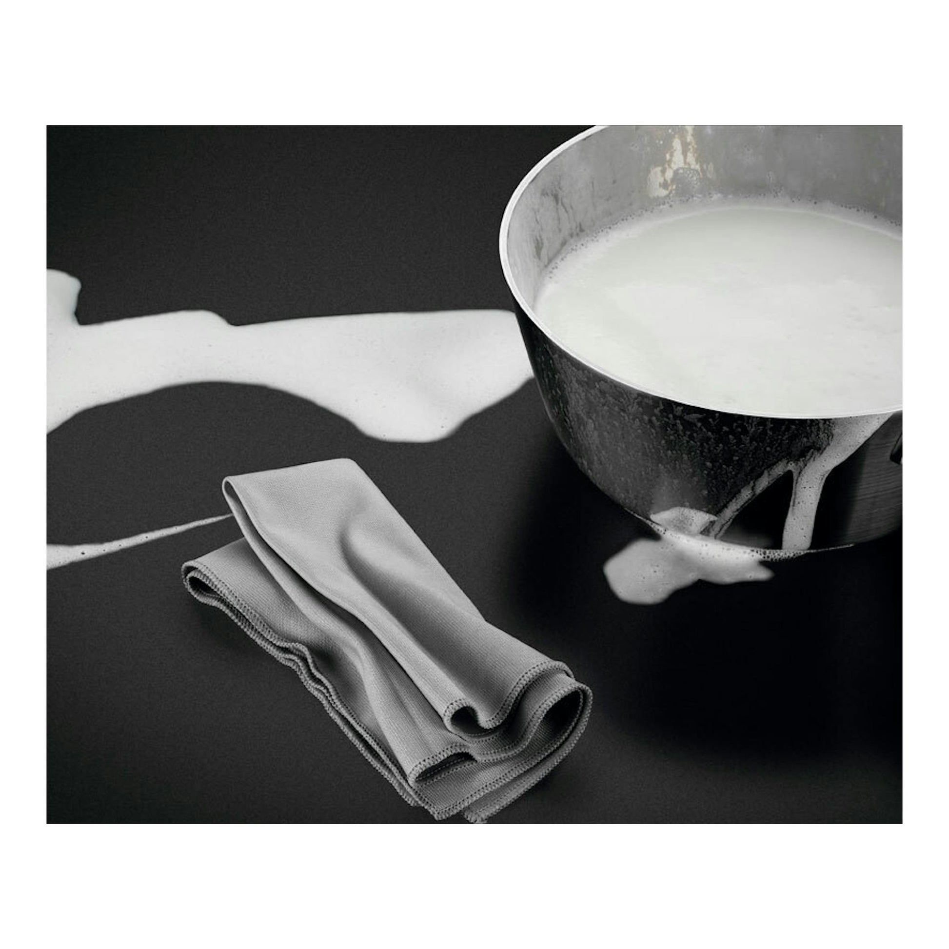 Placa de inducción negra con leche derramada, un paño doblado y una olla metálica.