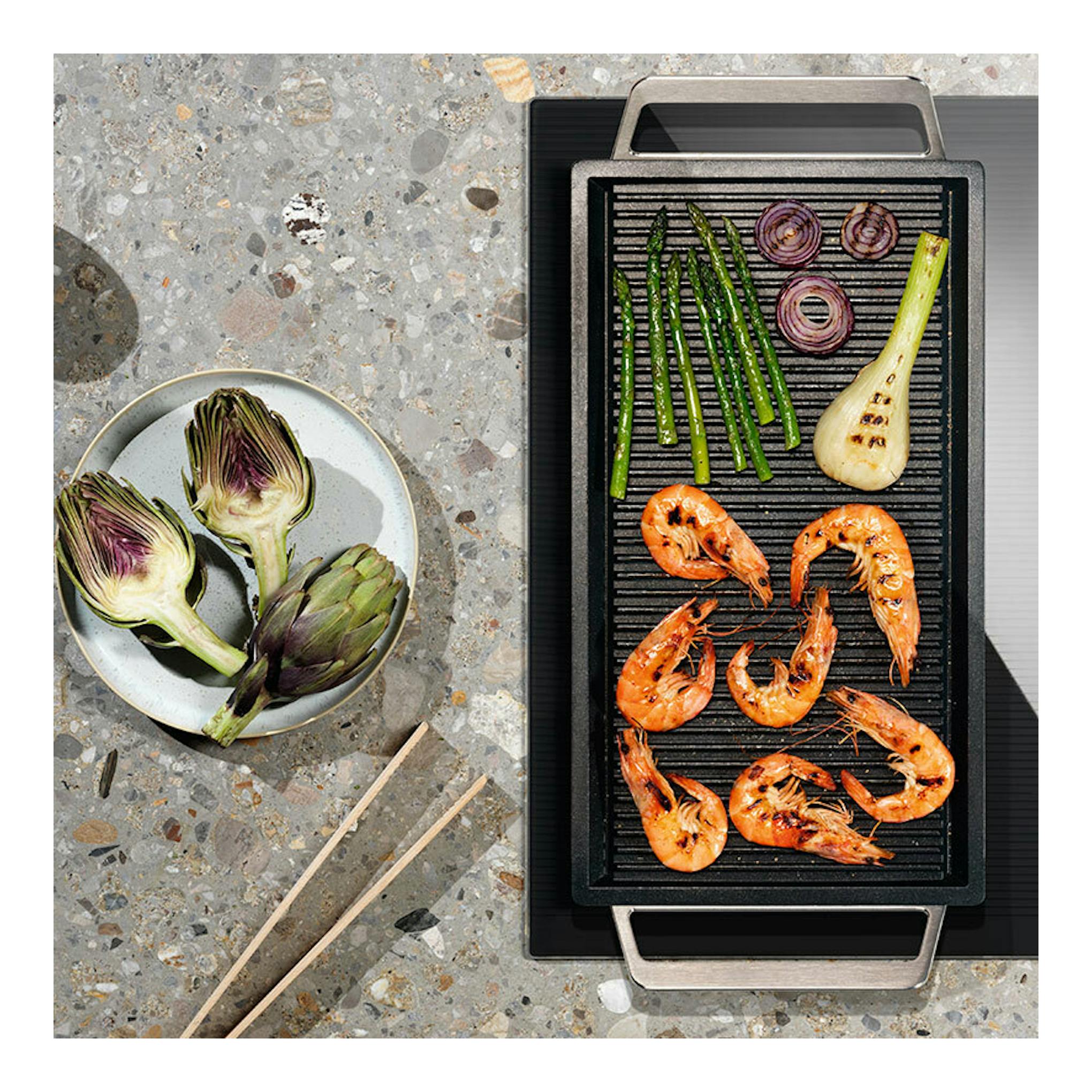 Placa de inducción Electrolux EIV633 de 60 cm, negra, vista cenital, con sartén grill y verduras y gambas.