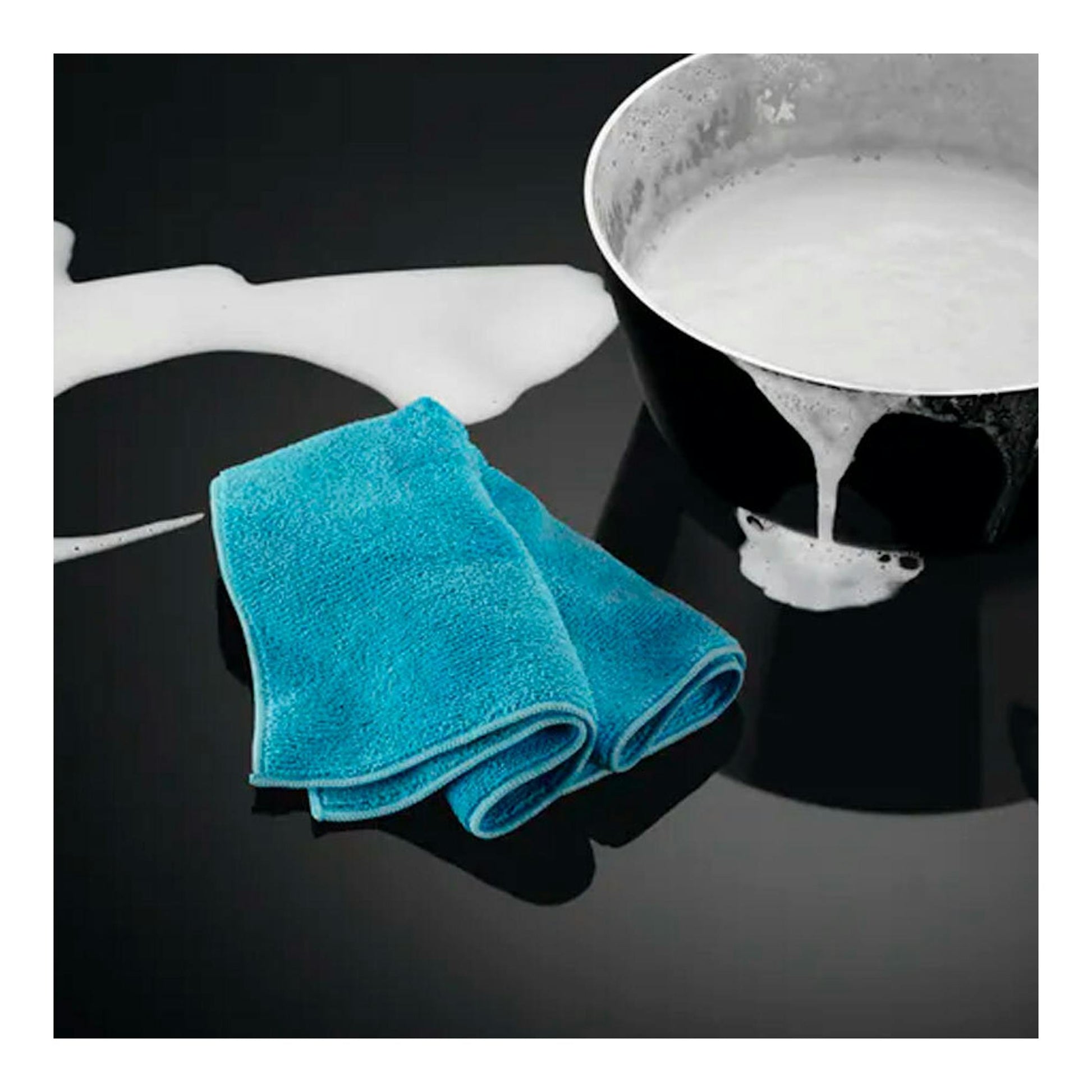 Placa de inducción negra en vista superior, con un cazo derramando leche y dos paños azules encima.